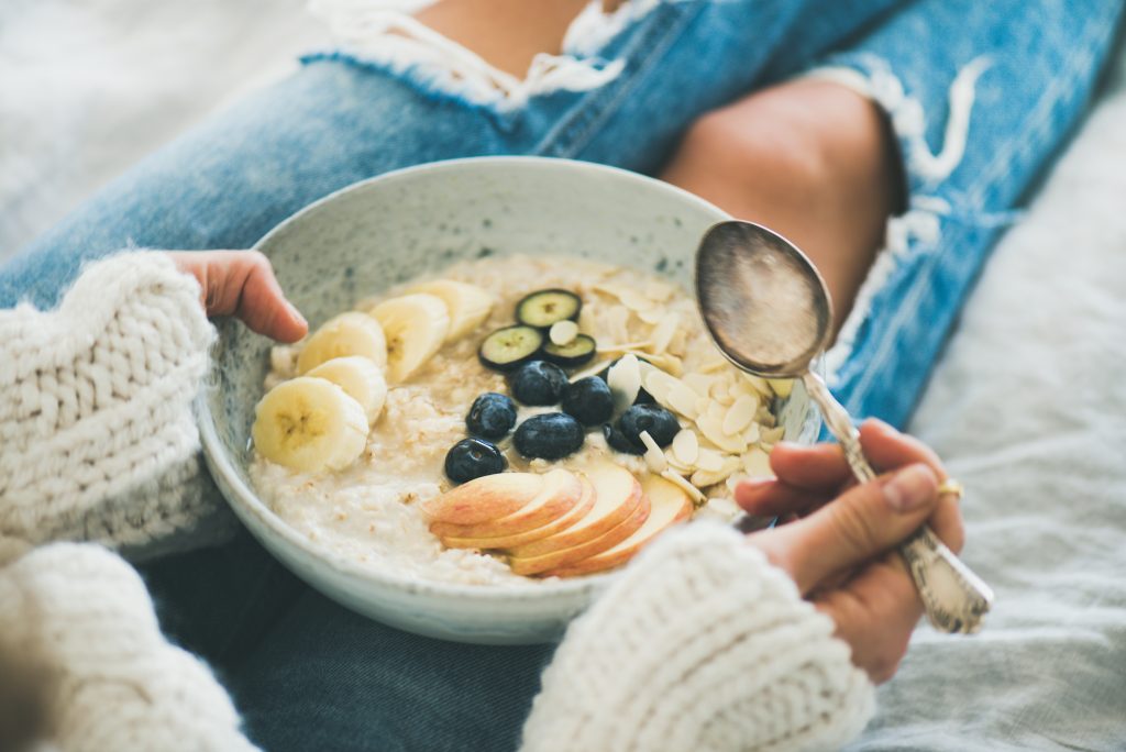 Gesundes Winterfrühstück im Bett. Frau in Wollpullover und schäbigen Jeans essen vegane Mandelmilch Haferflocken Brei in Schüssel mit Beeren, Obst und Mandeln. Clean Eating, vegetarisches Ernährungskonzept