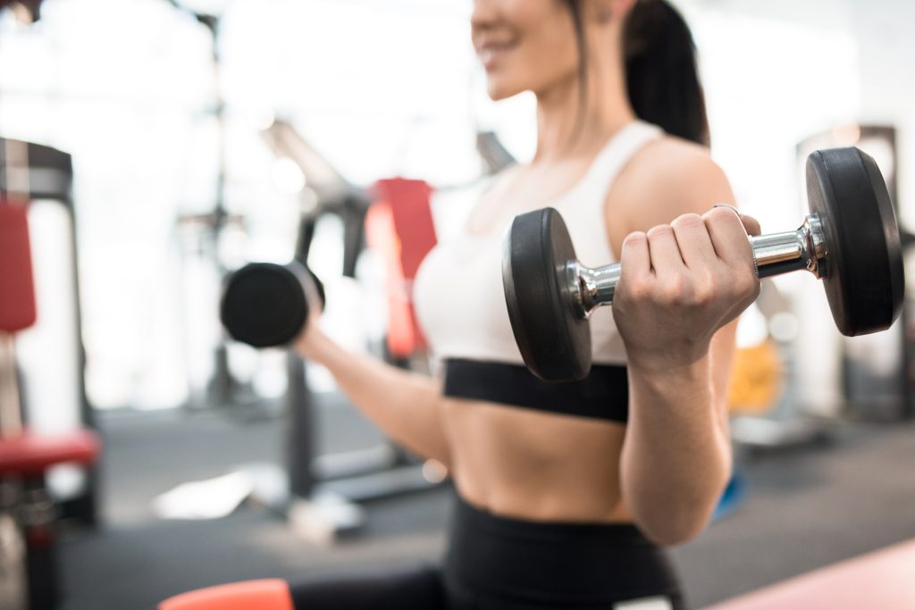 Nahaufnahme einer sportlichen jungen Frau, die in einem modernen Fitnessstudio mit Hanteln trainiert, Fokus auf schwere Hantel in der Hand